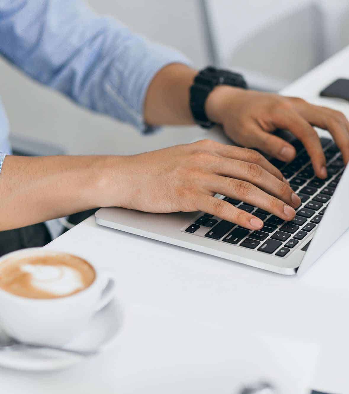 Man in blue shirt using laptop for work, typing on keyboard. Indoor portrait of male hands on computer and cup of coffee on table..