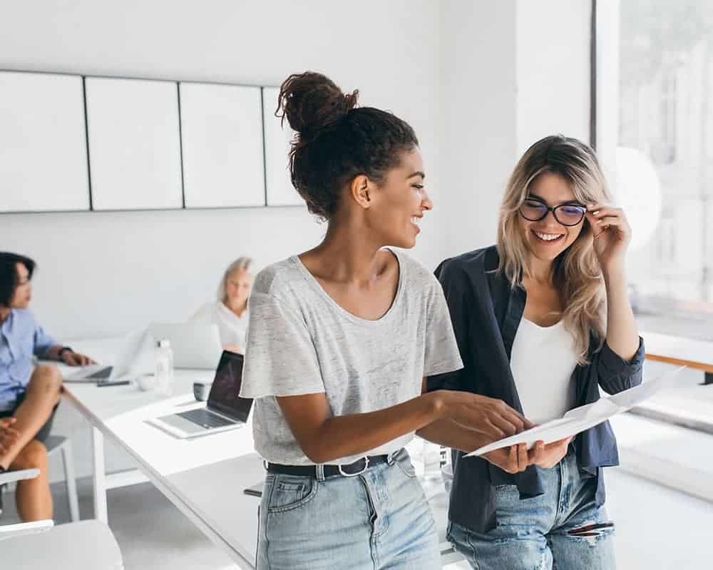 Young female executive explaines new strategy to blonde employee in glasses and smiling. Indoor portrait of multicultural collective working on project in office and using laptop..