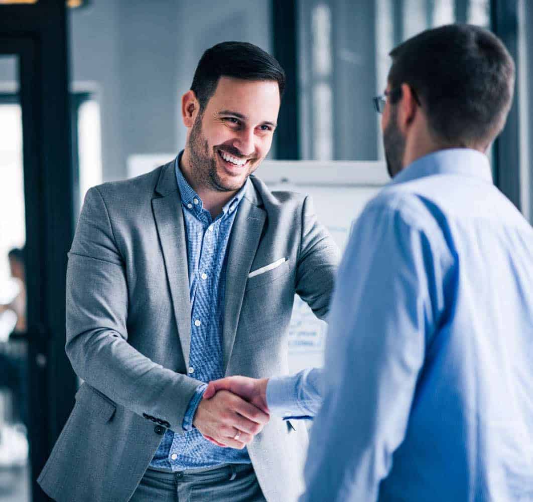 two-smiling-businessmen-shaking-hands-while-standing-office-min