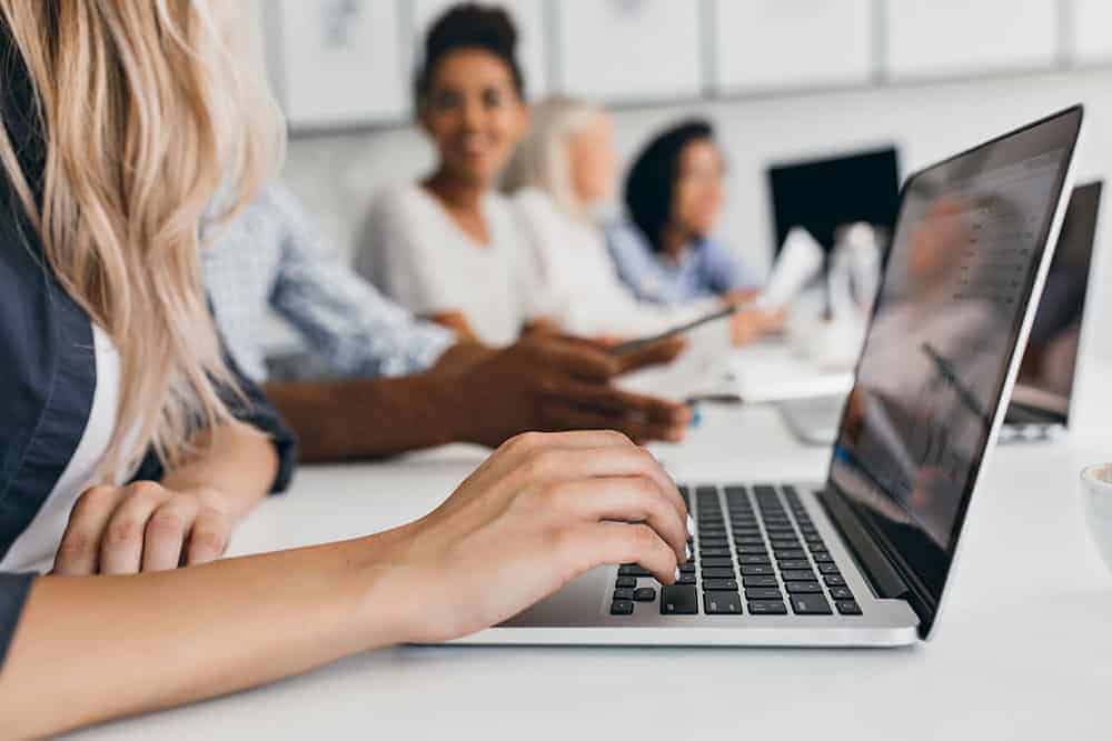 Blonde woman with elegant hairstyle typing text on keyboard in office. Indoor portrait of international employees with secretary using laptop on foreground..